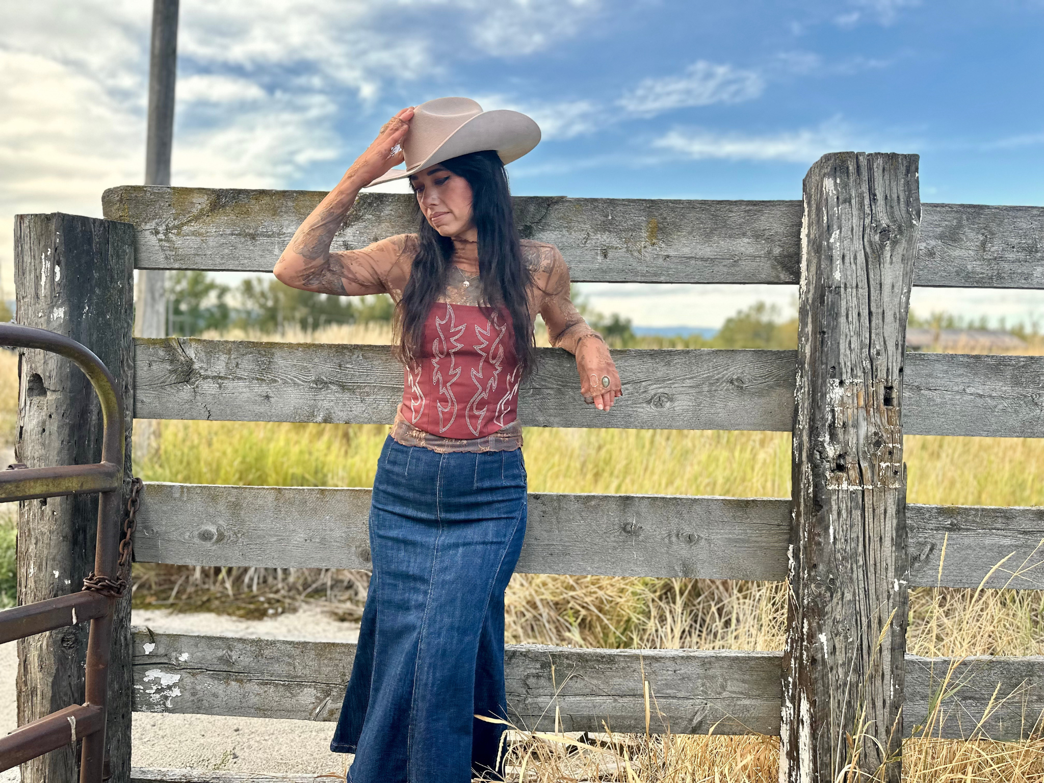 Woman in cowboy hat and patterned top leaning against a wooden fence in a rural setting